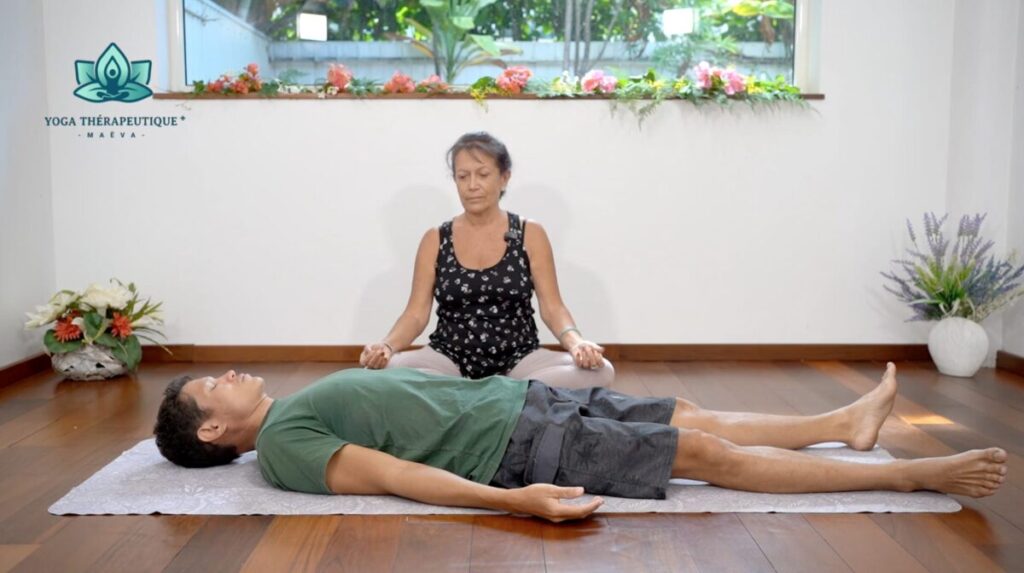 Une professeur de yoga assise guide un pratiquant couché devant elle.